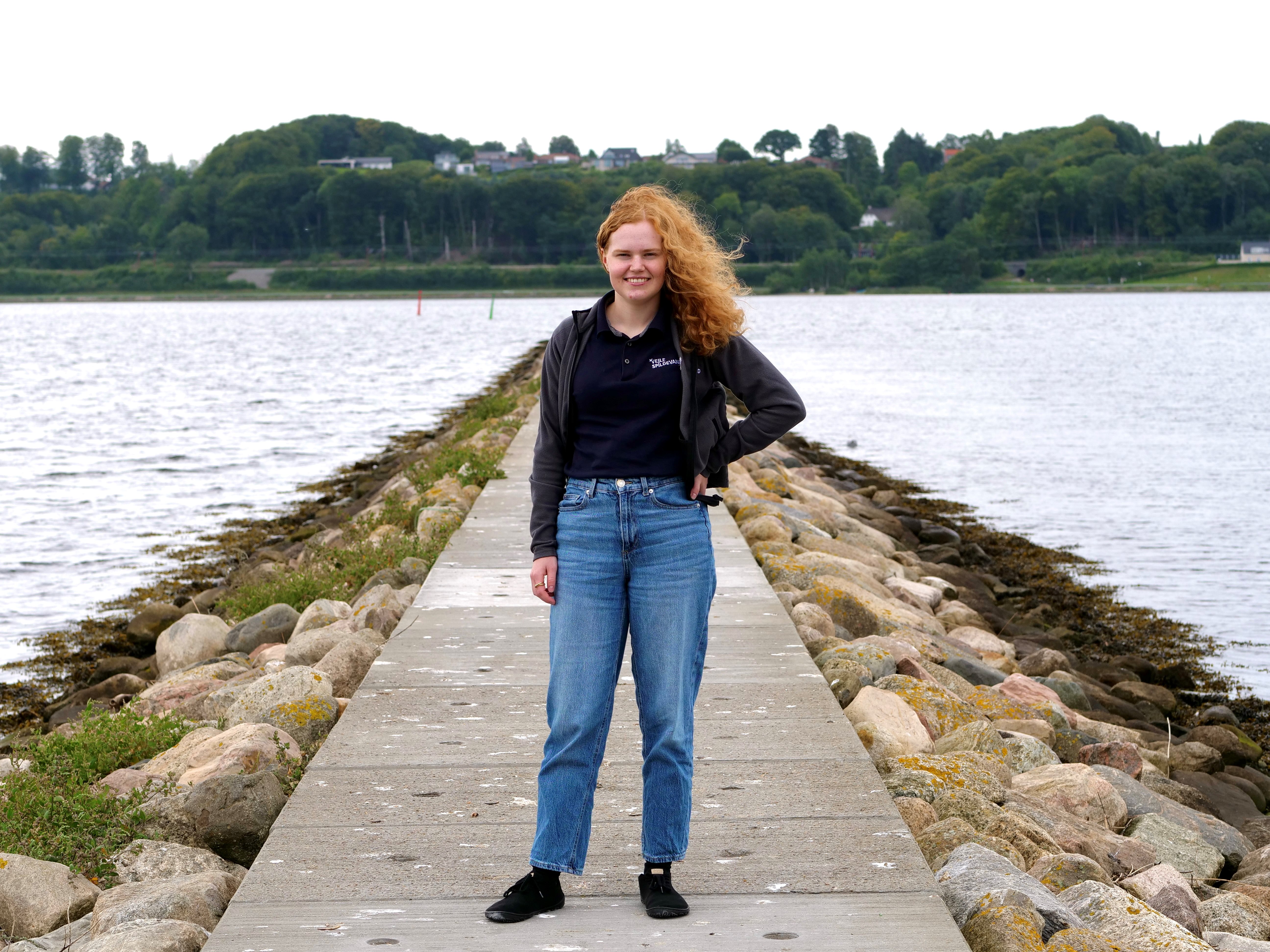 2025 10 02 En Ung Kvinde, Johanne Kaysen Thomsen, Omgivet Af Vand På Begge Sider. Hun Har Rødt, Krøllet Hår, Blå Jeans, Sort Trøje Og Mørke Sko Mads Volquartz