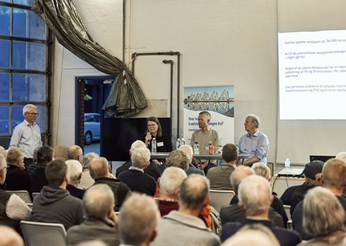 2026 09 30 Deltagere Sidder Tæt I Vejle Spildevands Garage, En Kvinde Taler I Mikrofon Ved Et Vejle Spildevand